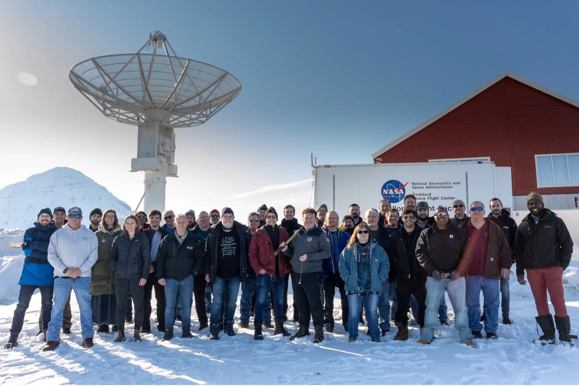Photo of 35 men and women standing in front of a satellite dish and truck with the NASA logo on it. there is snow on the ground a mountain in the background.