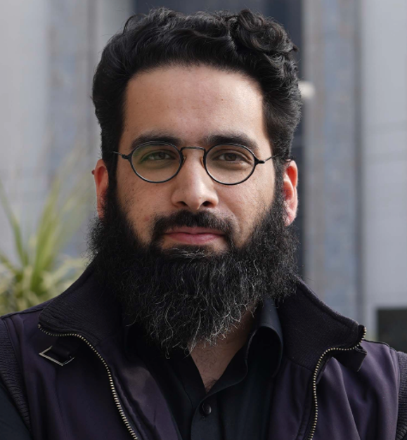 Portrait photo of a dark bearded man wearing dark clothing and glasses