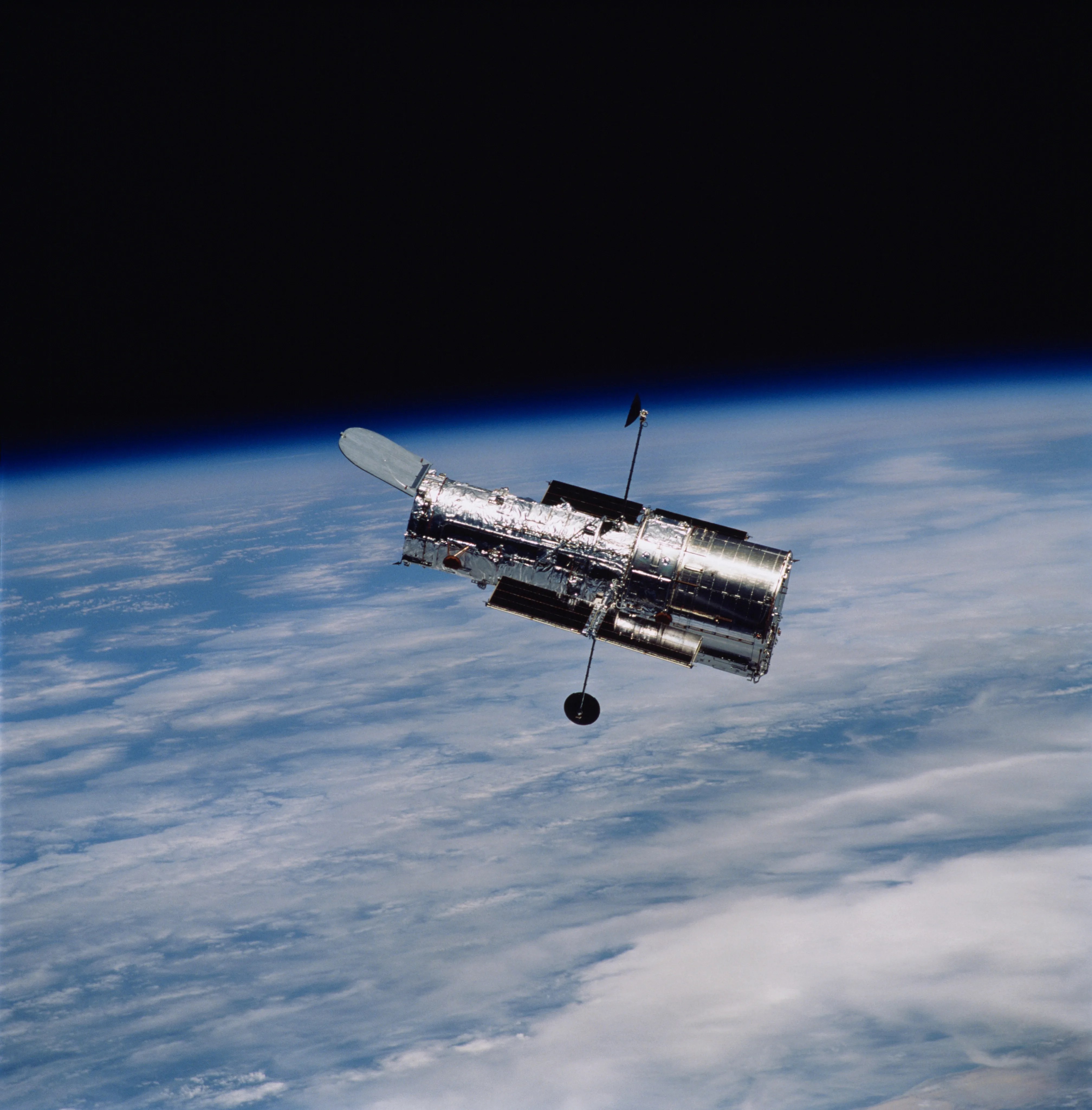 The Hubble Space Telescope is seen in orbit, with the cloudy and blue surface of Earth behind it, with black space above it.