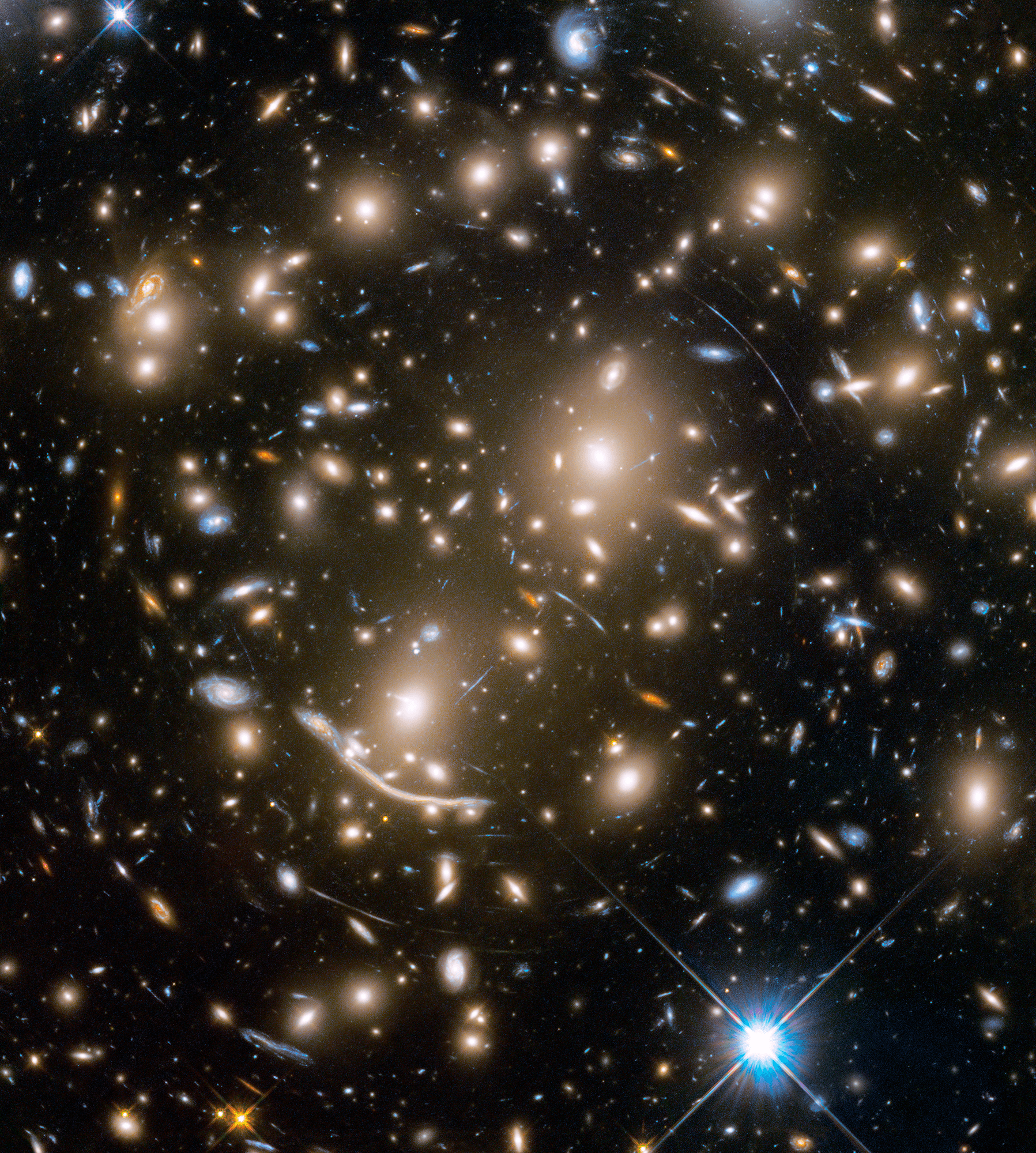 A packed field of galaxies and curved blue streaks and arcs. Bright blue-white foreground star at bottom right.