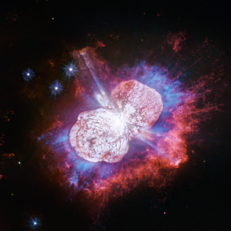 This image shows an exploding star. Two prominent lobes of white material expand outward from the center, surrounded by blue, pink, and red gas and dust against black space.