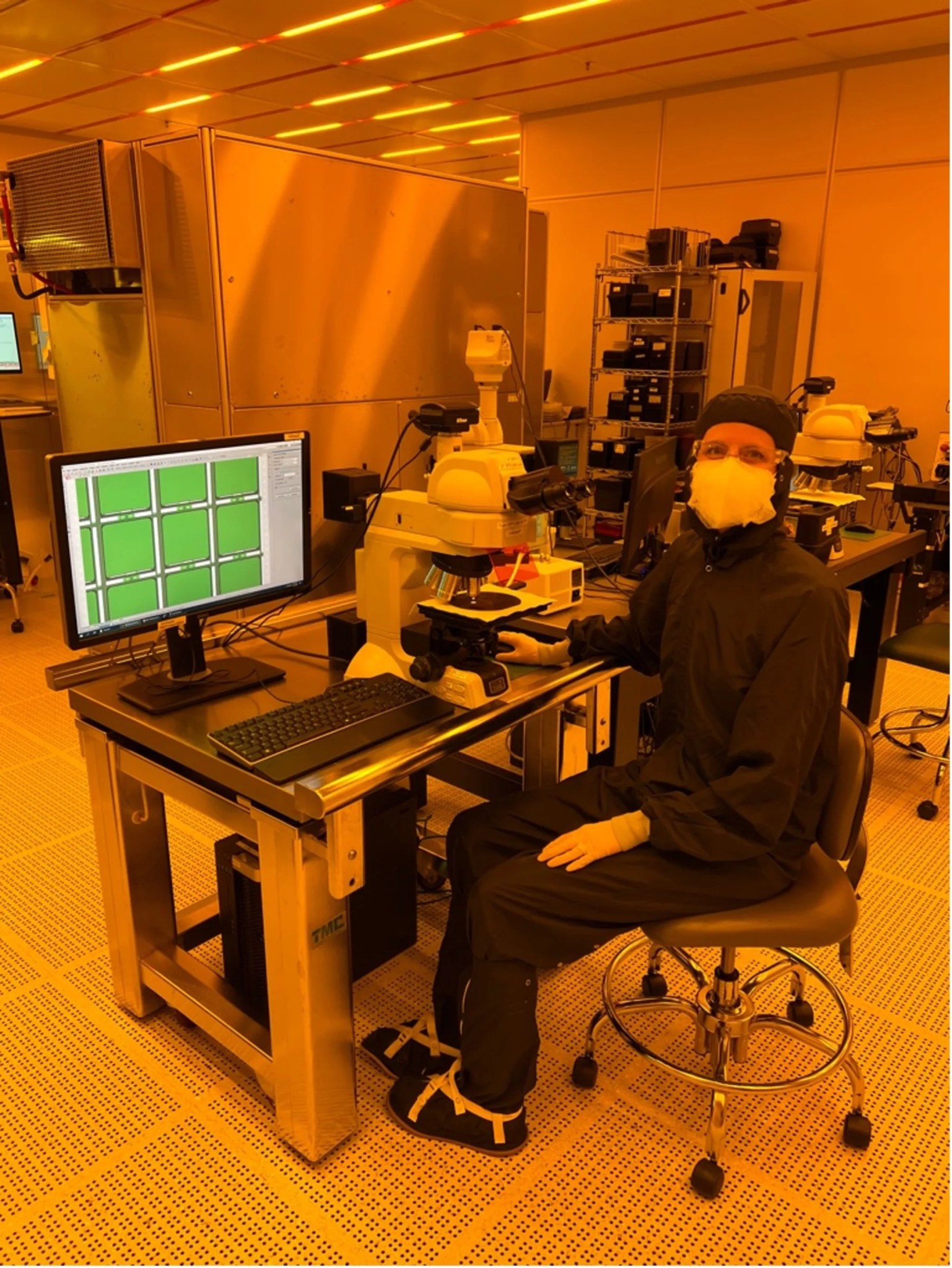 Photo of a woman in protective lab clothing sitting at a computer in a cleanroom with 9 green squares on the screen