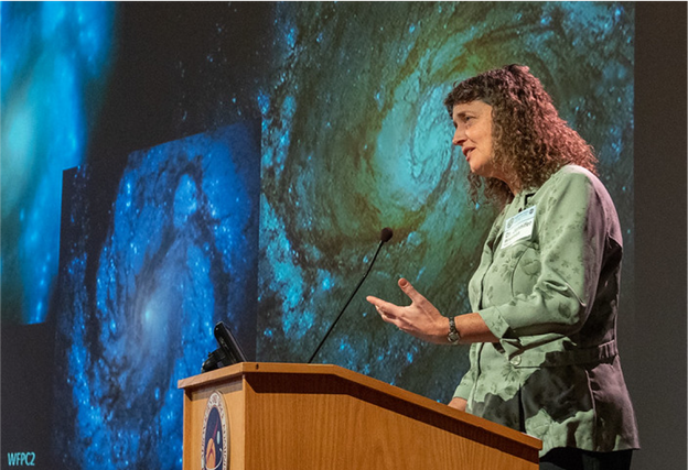 Jennifer Wiseman speaks to an audience from a podium. The project behind her show several swirling galaxies.