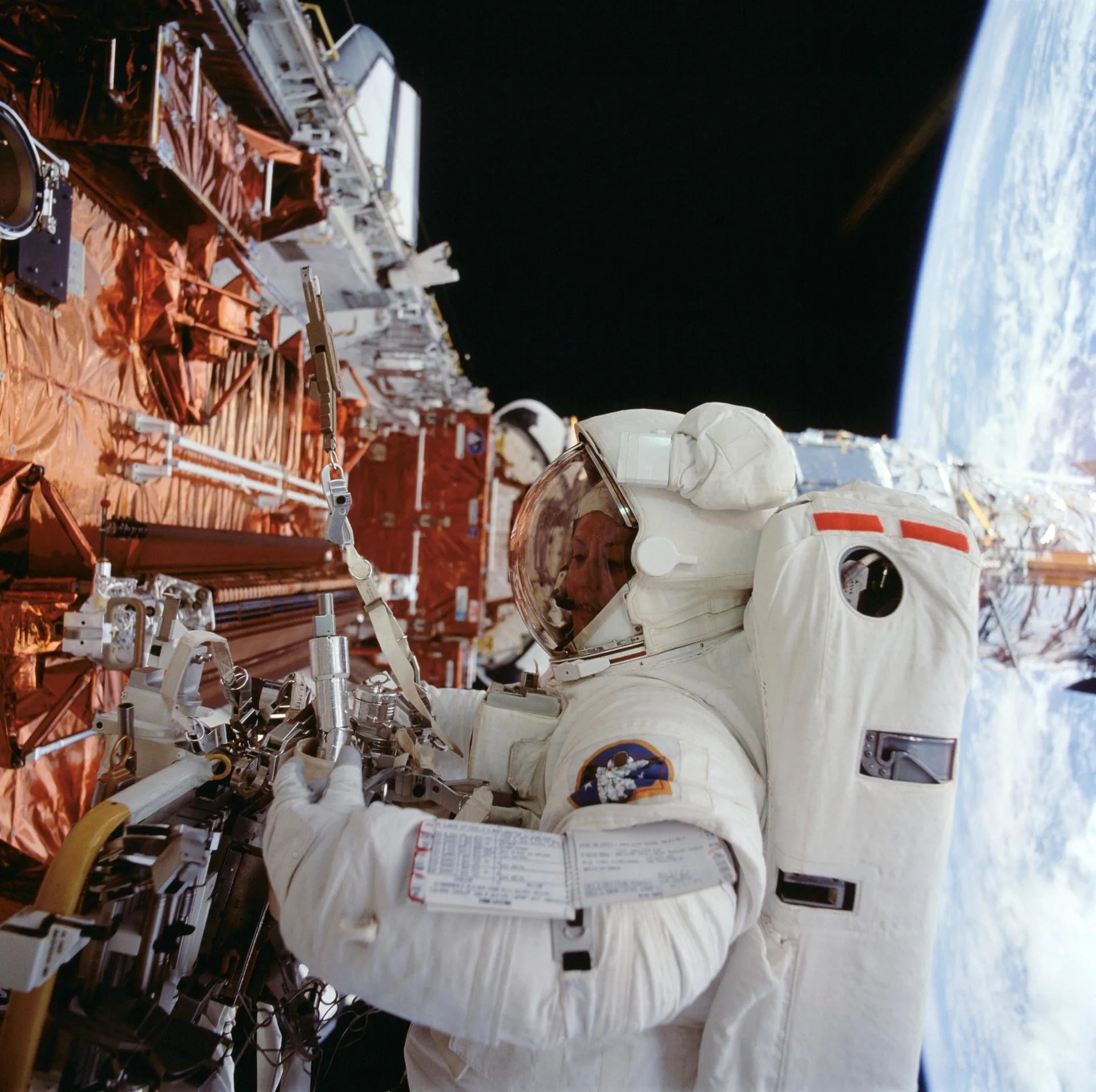 Astronaut Kathryn C. Thornton, center, grips a tool to perform servicing mission tasks on Hubble, left, with the Earth in the background on the right.