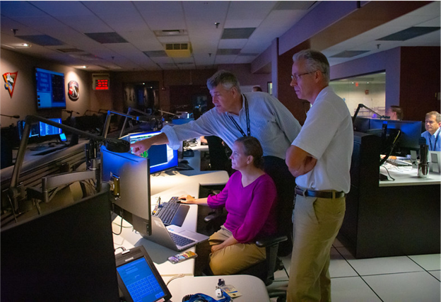 A woman (Morgan Van Arsdall) and two men (Larry Dunham and Joe Stock) are gathered around computers in the Operations Control Center, with more computers and staff in the background.