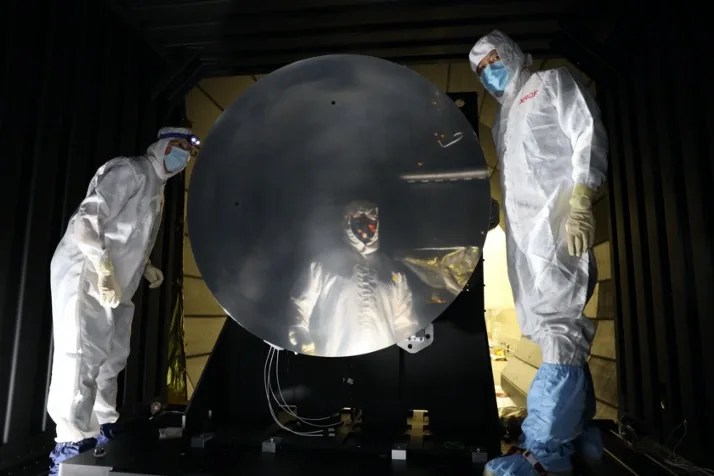 Photo of two male scientists in lab suits next to a large scale mirror and one scientist reflected in the mirror.