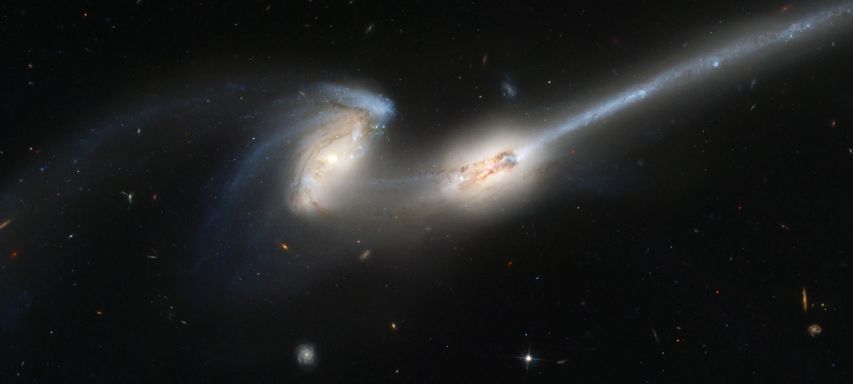 Two interacting galaxies centered in the image. The left galaxy is a spiral galaxy with its arms distorted and drawn out toward the left side of the image. The right galaxy has a long tail that extends to the upper-right corner of the image. Gas and dust also extends between the two galaxies.