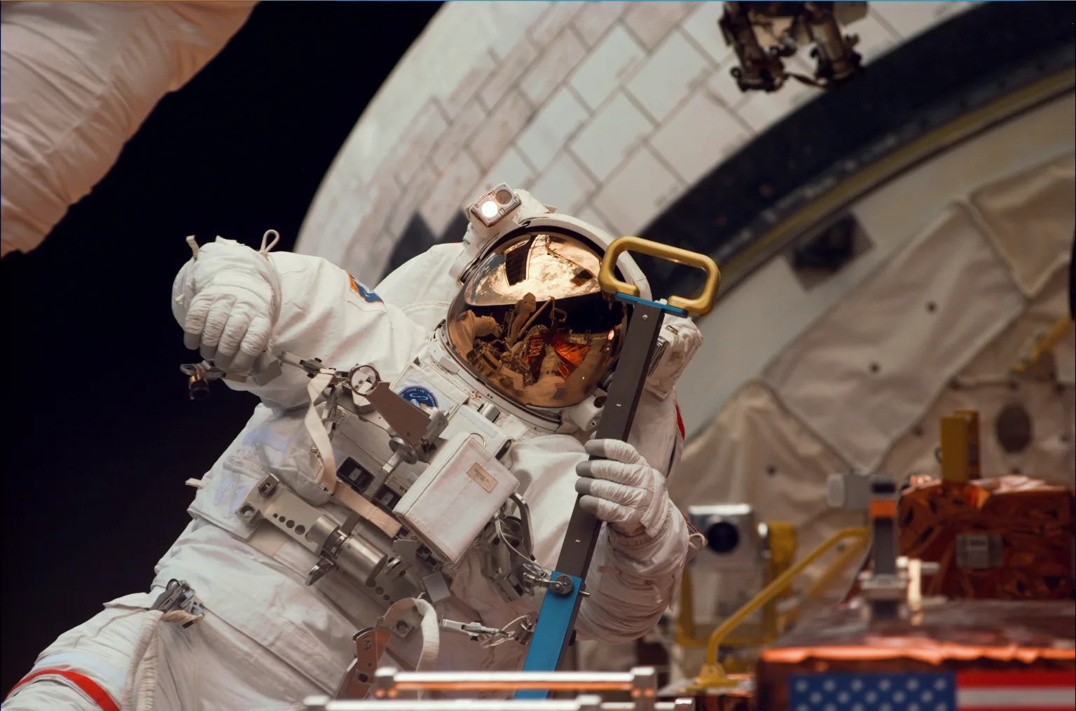 Astronaut in a white space suit in space doing maintenance on the Hubble Space Telescope with the shuttle Discovery's cargo bay door open.