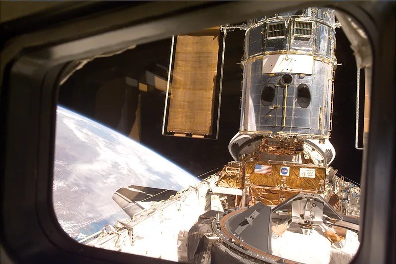 View from out of a shuttle window of the Hubble Space Telescope in space with the Earth in the background.