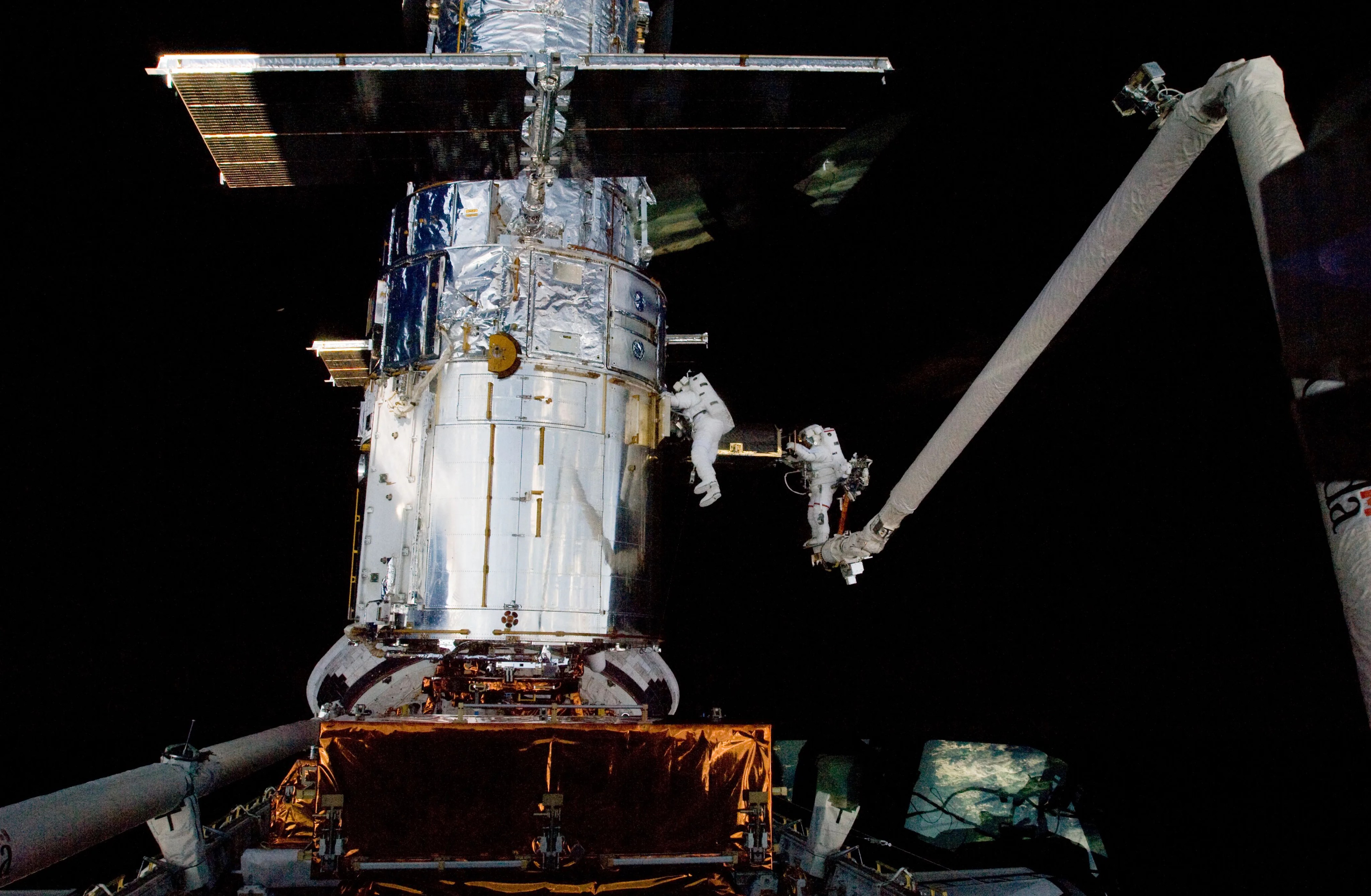 Two astronauts in white space suits tethered on a robotic arm servicing the Hubble Space Telescope in space.