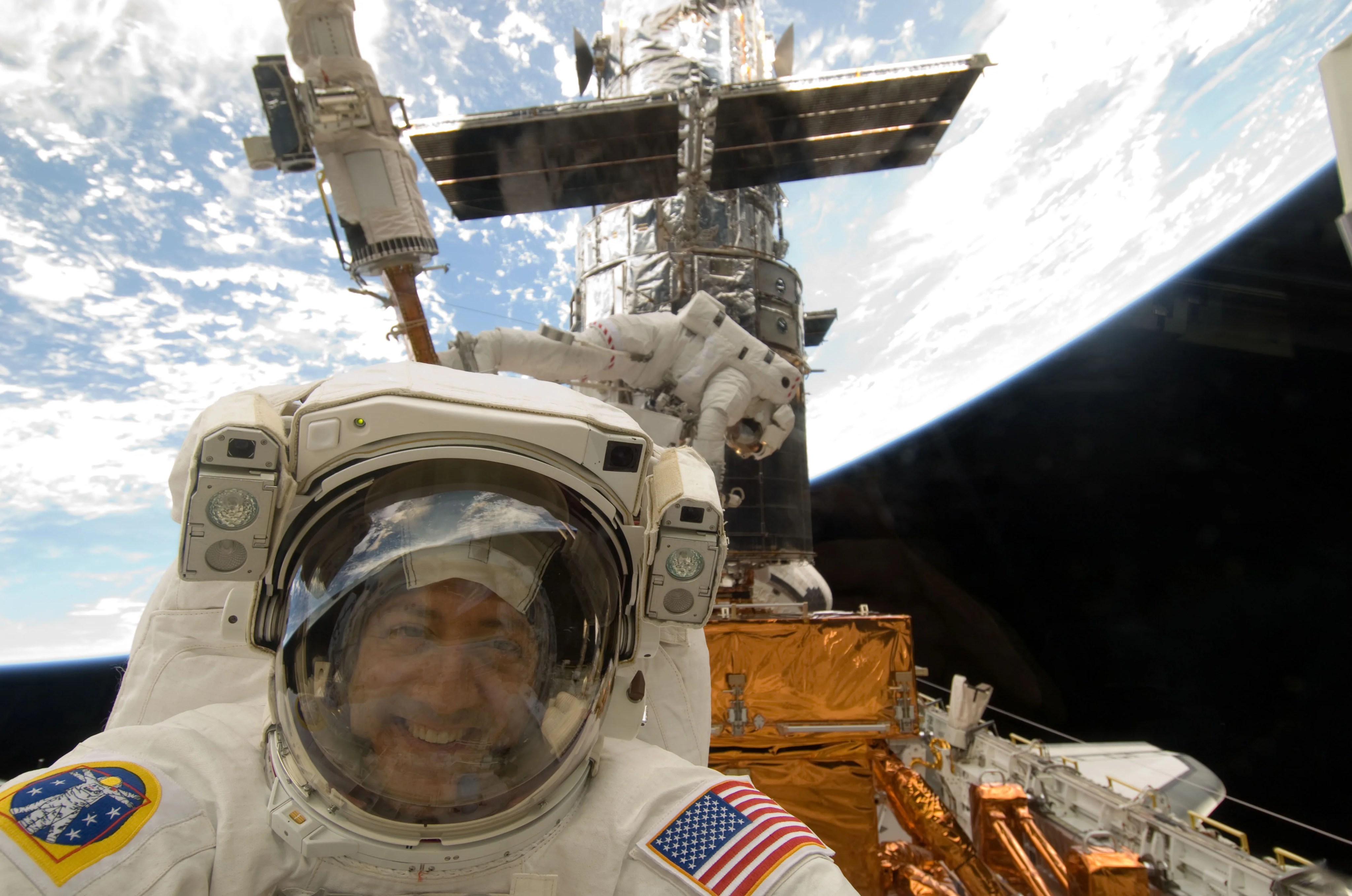 Astronaut in a white space suit smiling in a selfie with a crewmate, the Hubble Space Telescope and the Earth in the background.