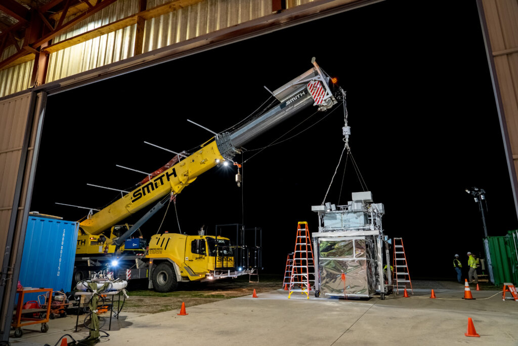 The EUSO-2 payload hangs from a yellow crane vehicle. The payload main body is mostly enclosed silver plastic wrapped wraps with multiple bars across the top with various instrumentation.