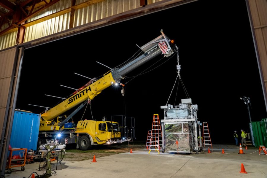 The EUSO-2 payload hangs from a yellow crane vehicle. The payload main body is mostly enclosed silver plastic wrapped wraps with multiple bars across the top with various instrumentation.