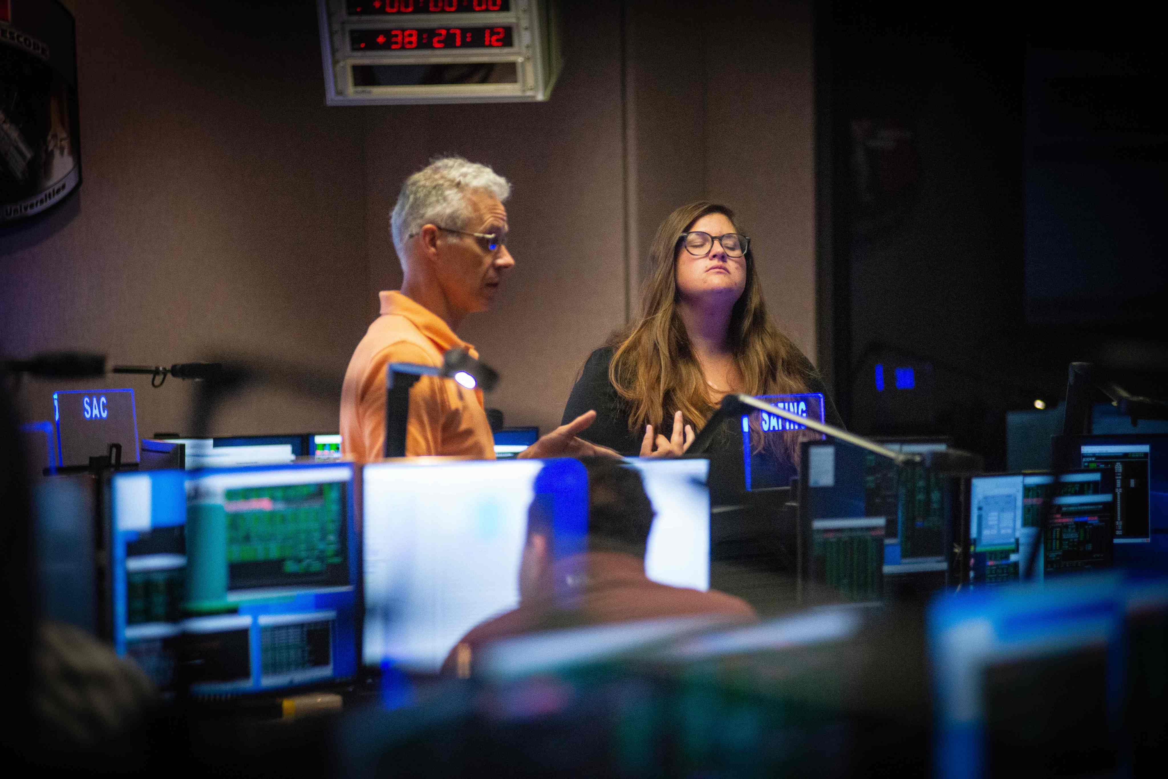 A man and a woman standing in the Hubble control center both look concerned regarding the process of switching to backup spacecraft hardware.