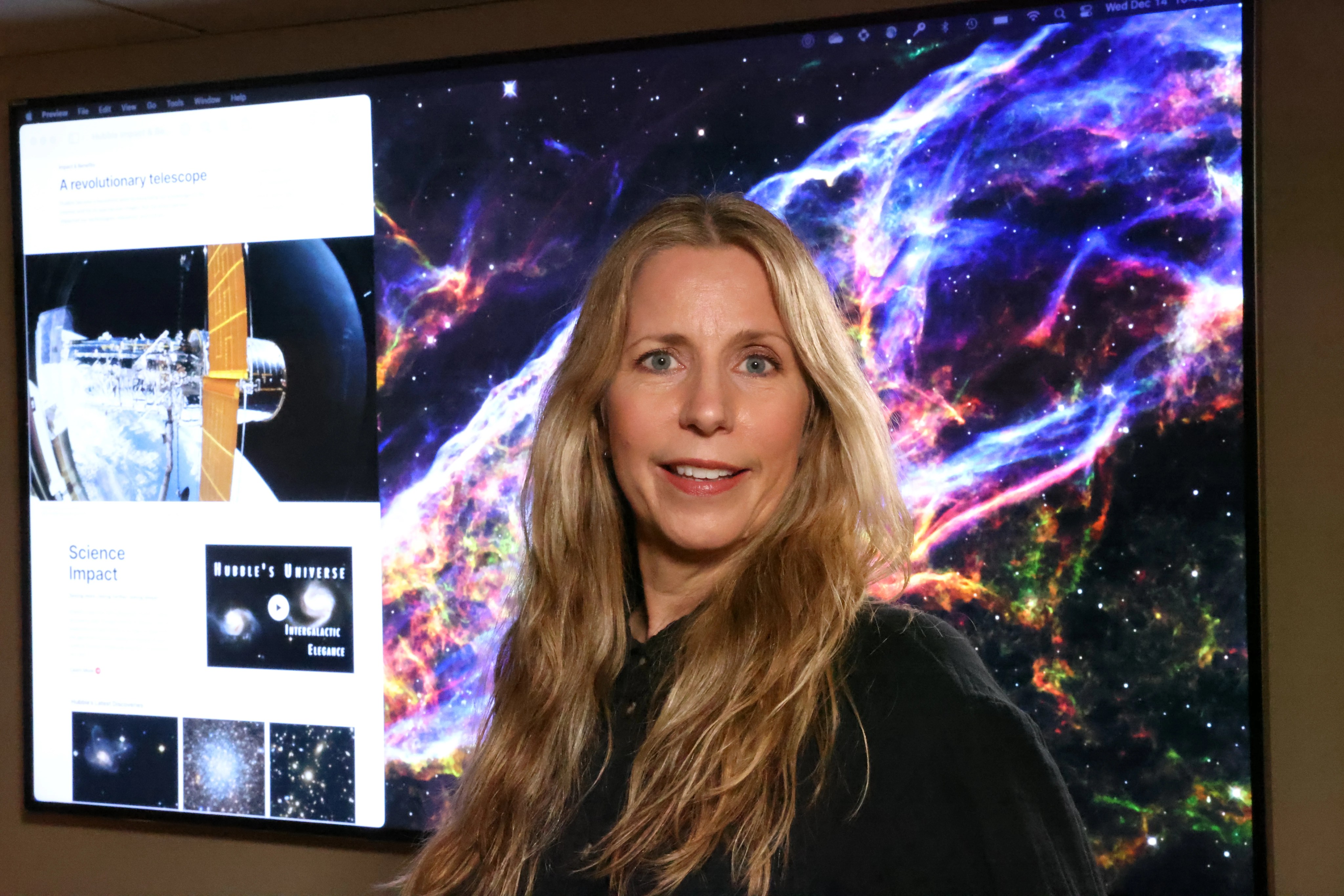 headshot of a woman with blonde hair in a black shirt standing in front of a hubble image on a tv monitor