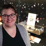 Headshot of a woman with glasses in a light gray sweater standing in front of the Hubble display case.