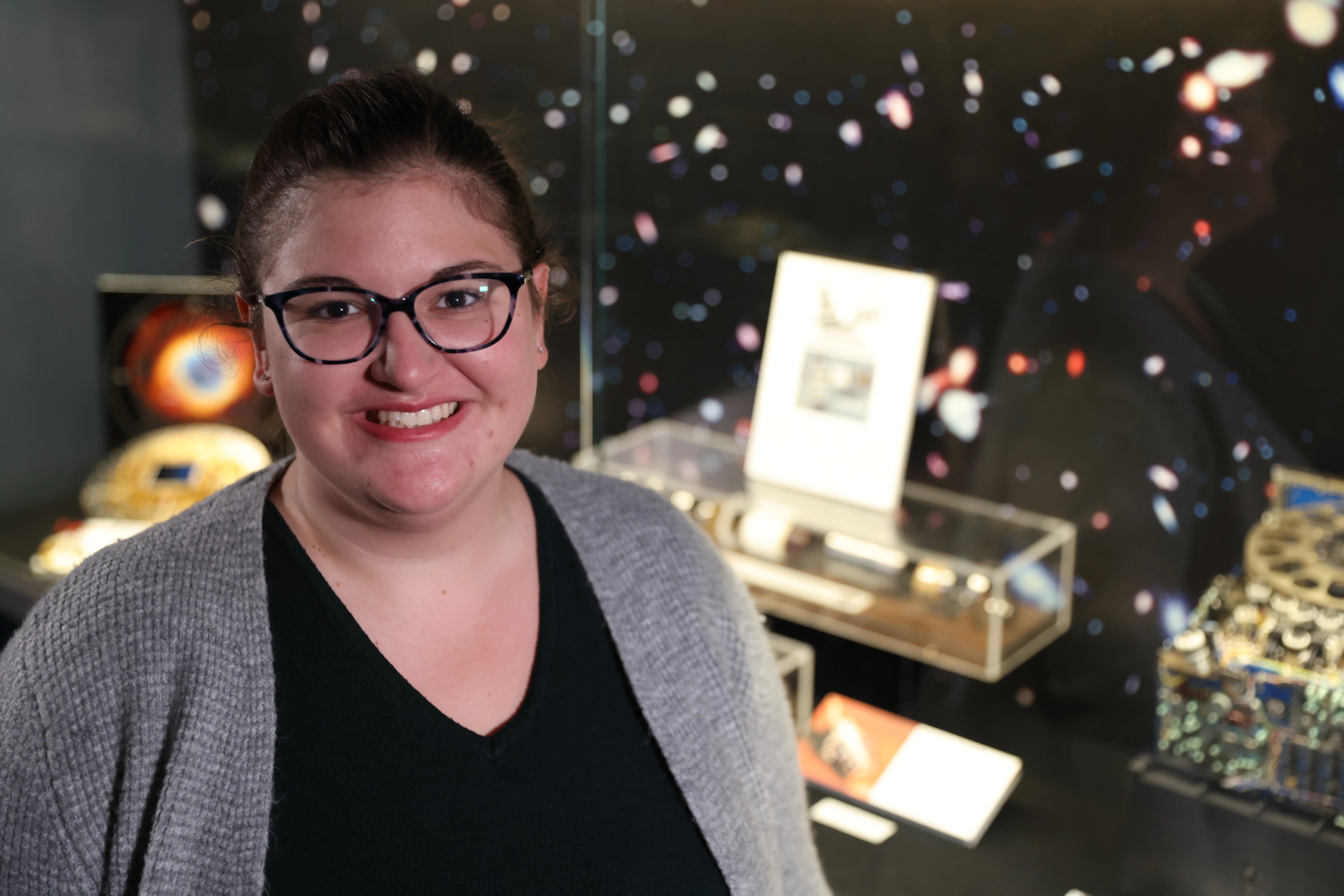 Headshot of a woman with glasses in a light gray sweater standing in front of the Hubble display case.
