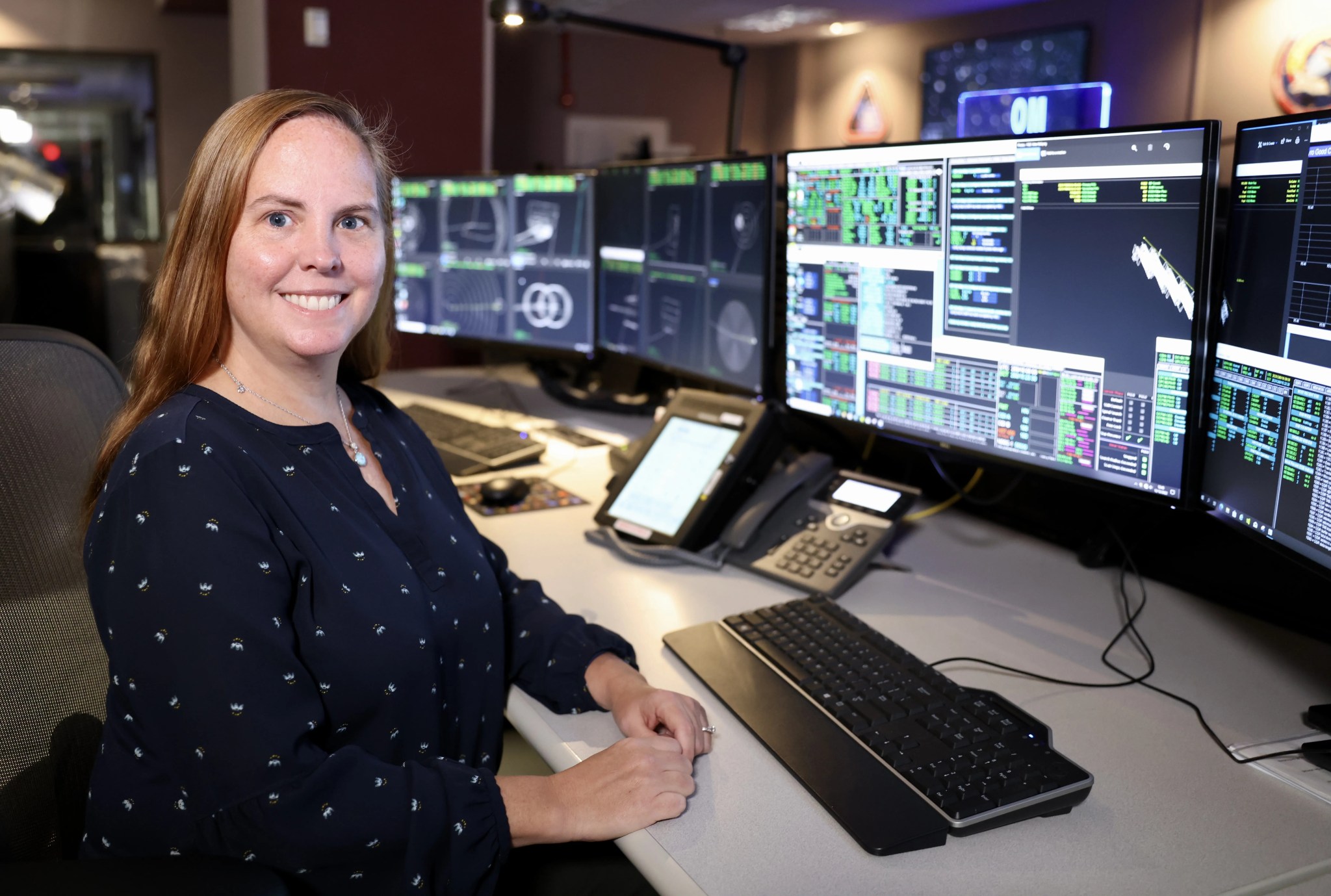 woman sitting at a computer desk in front of a bank of computer monitors, a phone and a keyboard