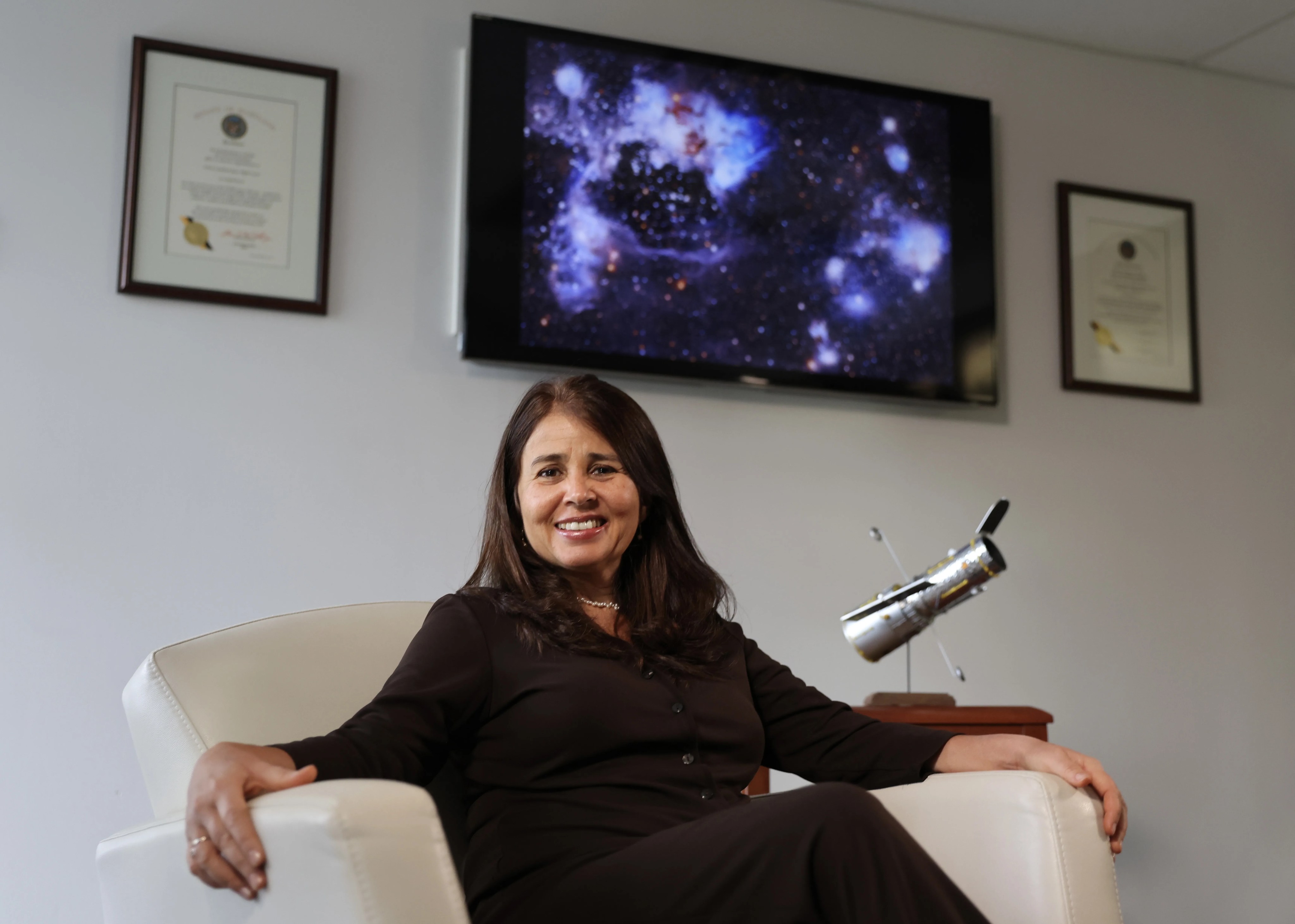 headshot of a woman wearing a dark brown sweater and brown hair sitting a white chair with a Hubble spacecraft model on the right