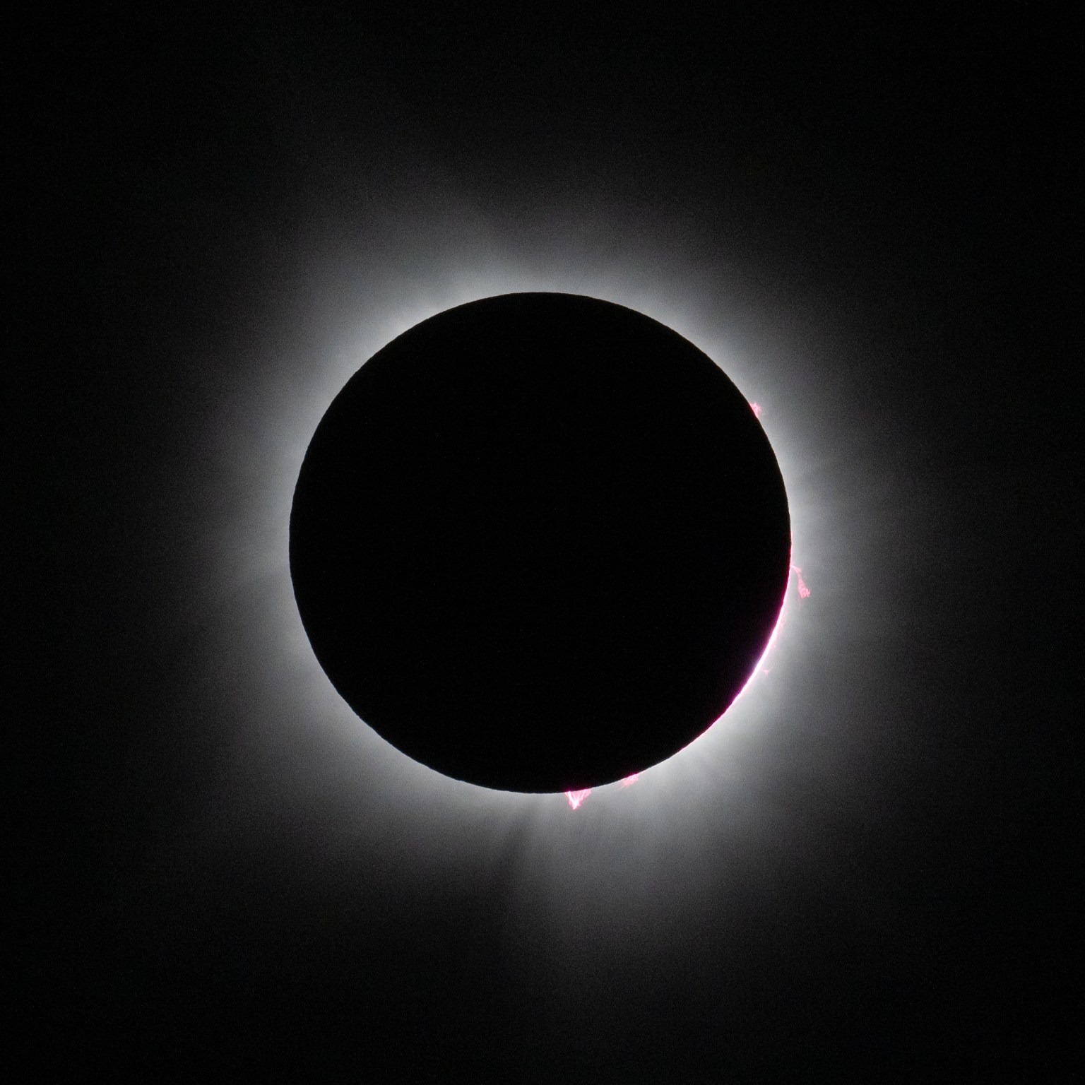 A large black disk is in the center. This is the Moon. Around it, white streams of light glow against a black background. On the bottom of the disk and on the right side, there are small, bright pink triangles of light.