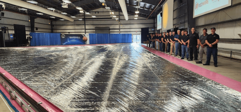 Photograph of 17 people standing to the right of a large shiny sail