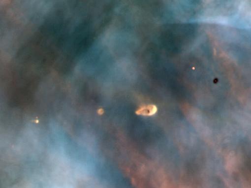 The view is filled with wisps of clouds in colors of white, grey, brown, and black. Protoplanetary disks appear as dark or bright points surrounded by denser white clouds. Center of the image holds a bright protoplanetary disk that is larger than the others.