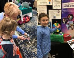 Left: Sharon Bowers demonstrates an app to a young girl. Right: Girl with her finger on a spinning multicolored wheel at an exhibition booth.