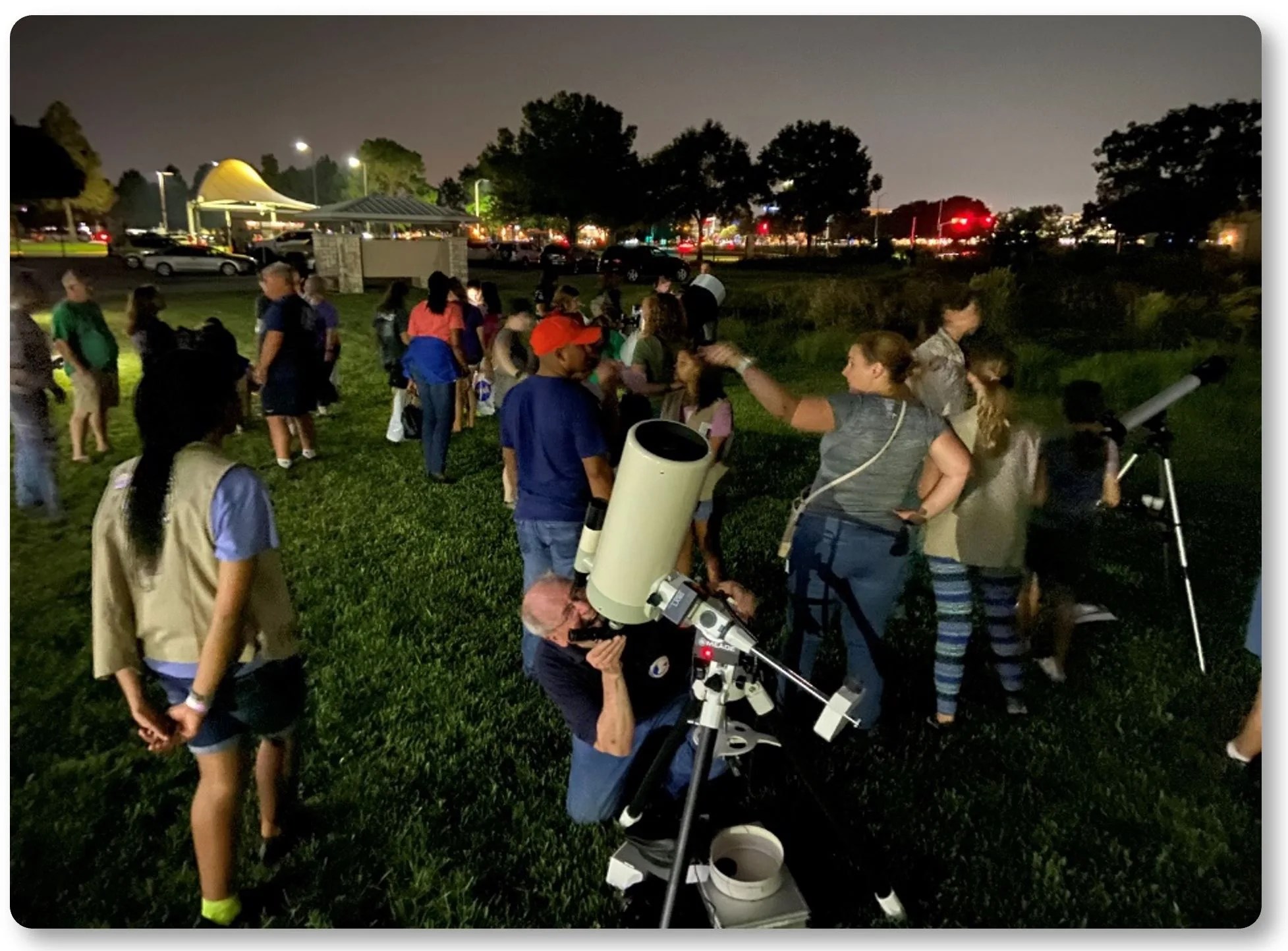 A man with grey hair looks through a telescope at night. He is crouched on the grass in a small crowd.