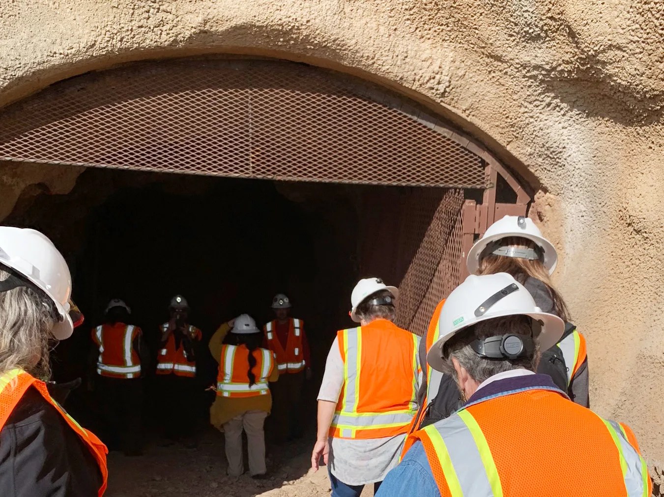 A small group of people wearing safety gear walk into a mine shaft.