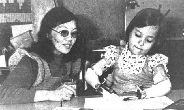 Black and white photo of a teacher with glasses seated at a table next to a young girl with prosthetic arms.