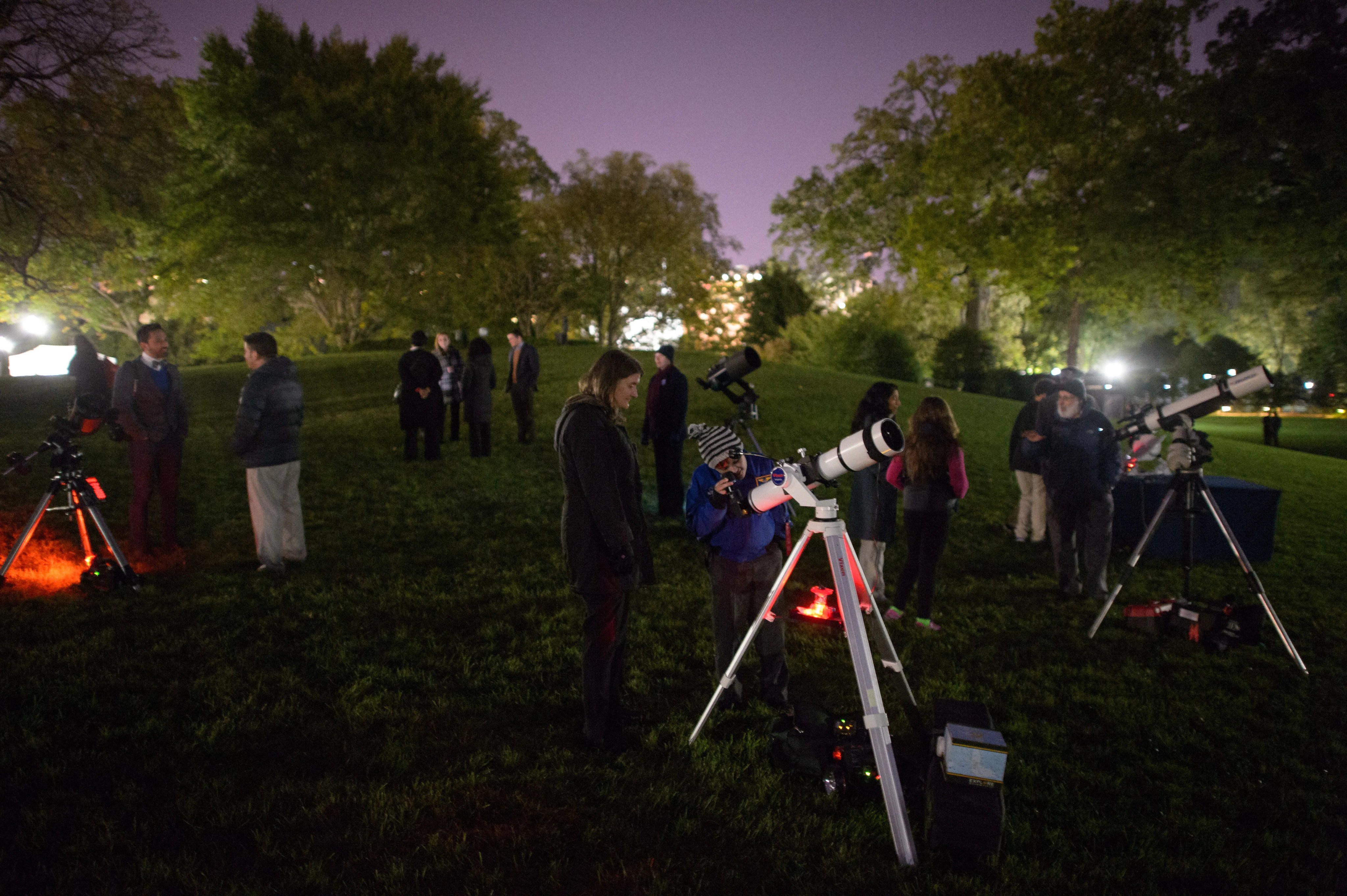 A child gazes into the eyepiece of a small telescope set up on a grassy lawn at night as an adult woman looks on. Other observers and telescopes are nearby.