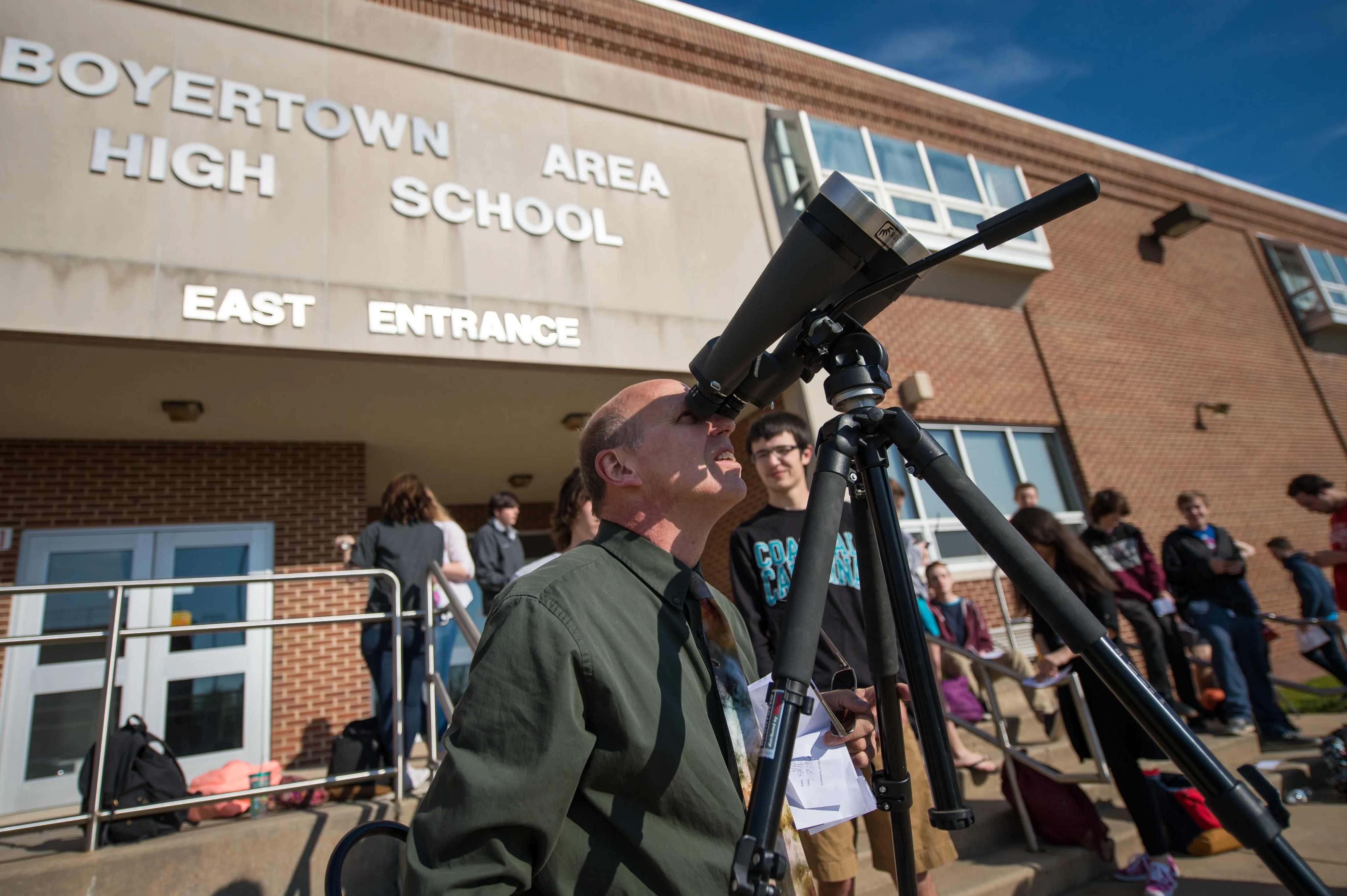 Outside a high school building, an adult man looks at the Sun through binoculars outfitted with a solar filter for safe viewing. About a dozen students are visible in the background.