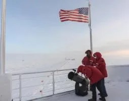 Three people in orange parkas with a telescope at the South Pole.