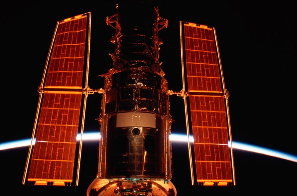 The Hubble Space Telescope is seen washed in gold hues from the sun, against black space and the curve of the Earth behind it.