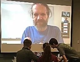 Three students in front of a projector screen and laptop. On the displays are a bearded man via webcam.
