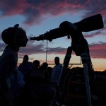 After sunset, with oranges and pinks in the darkening sky, a young person appears in silhouette, looking through a telescope, pointed toward right. A group of adults, also in silhouette, are visible in the background.