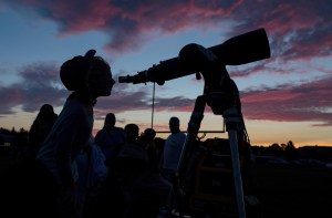 After sunset, with oranges and pinks in the darkening sky, a young person appears in silhouette, looking through a telescope, pointed toward right. A group of adults, also in silhouette, are visible in the background.