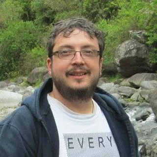 Portrait photo of a man wearing glasses and a hoodie and standing in front of a stream and green trees