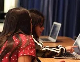 A young girl points at a laptop screen for another girl.