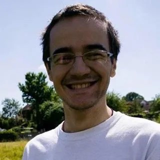 Photo of a smiling man wearing glasses and a tshirt and standing in a field outside