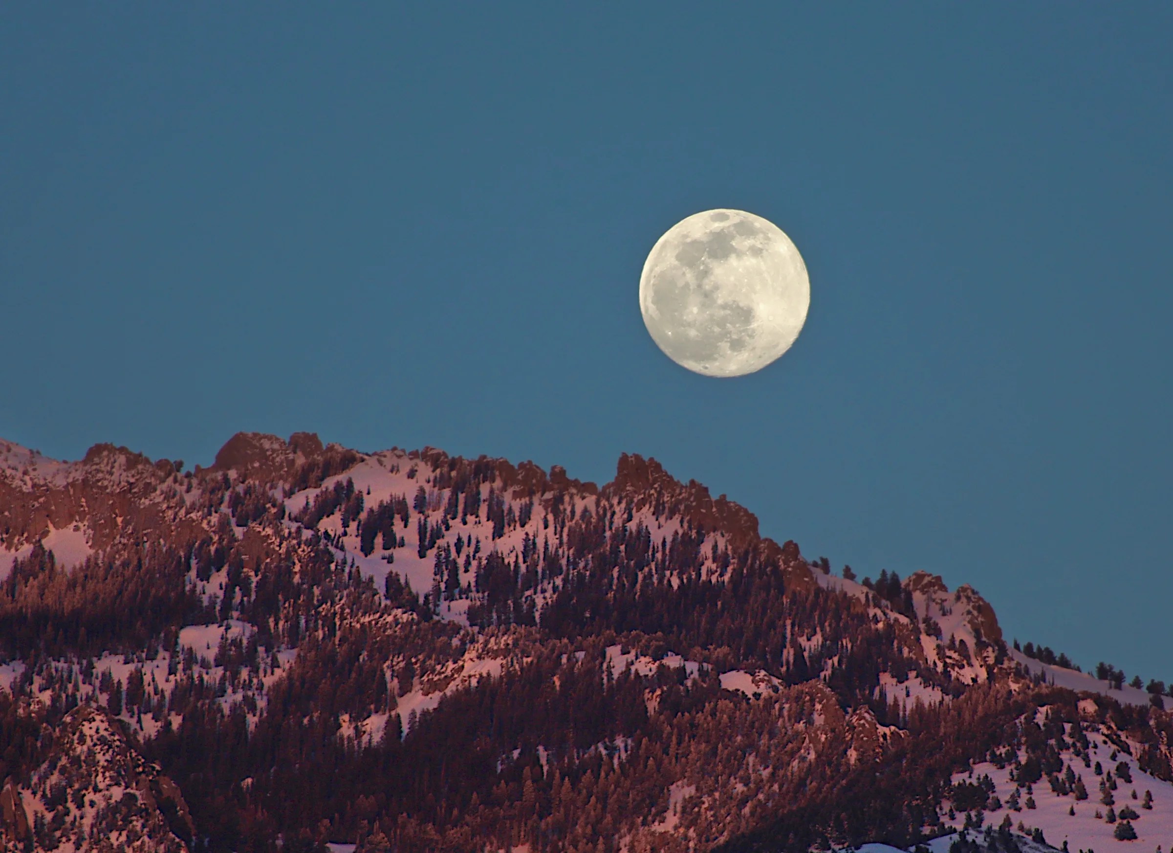 In a dark blue, evening sky, a bright, round full moon appears above a rugged, snow-covered mountain. Pink light from sunset colors the mountainside.