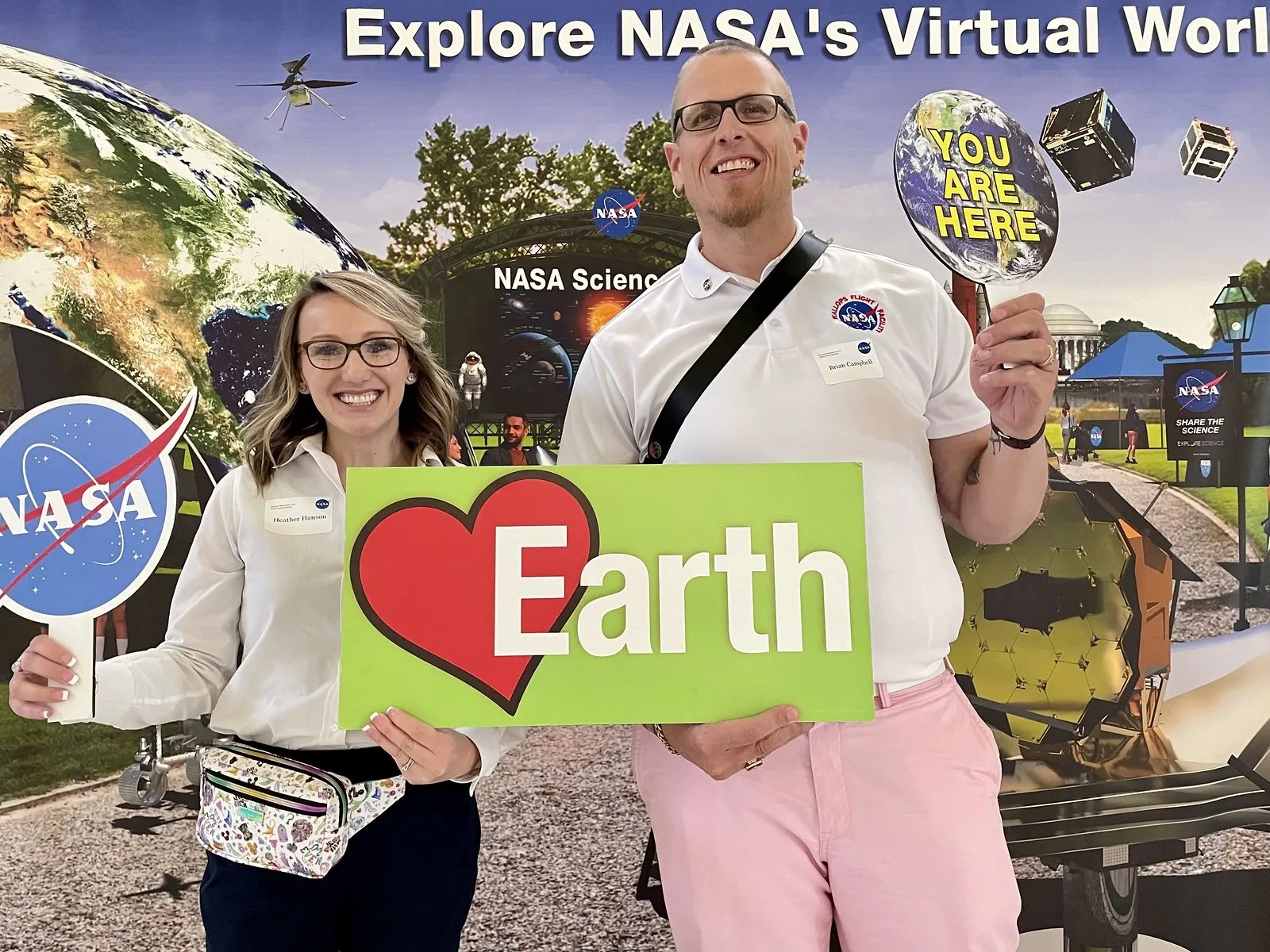 Photo of a smiling man and woman holding NASA signs and insignia and an Earth poster with a red heart.
