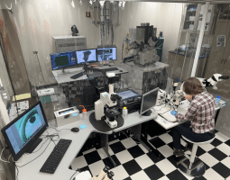 This image shows various microscopes available for use at the Advanced Electron Microscopy Center. An individual is utilizing one of the microscopes in the foreground and data from another instrument is portrayed on various computer screens in the background.