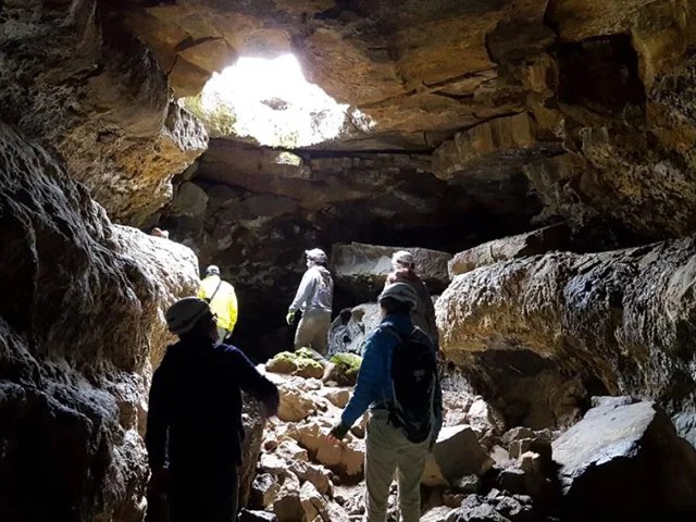 Five humans explore an open cave. Light from the Sun filters in through a hole at the top.