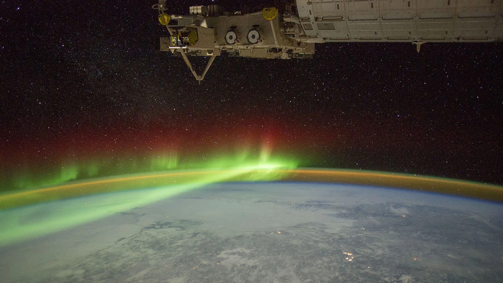 The limb of Earth with glowing auroras and a thin band of atmosphere under part of the International Space Station.