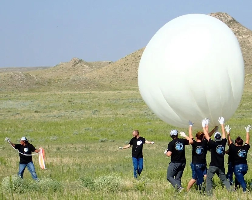 Photo of a group of 7 people in a field wearing black shirts and pants and handling a large white weather balloon