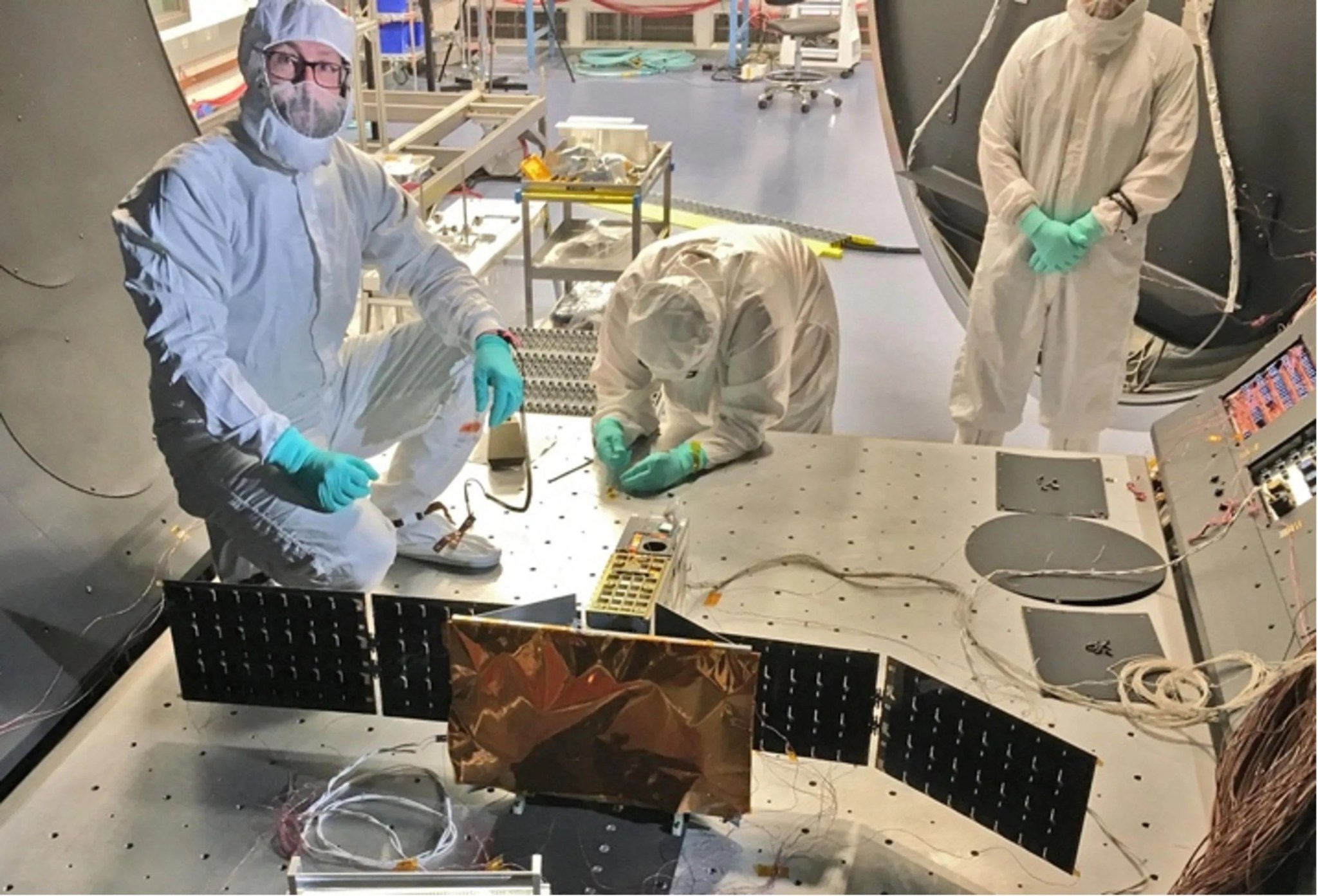 Photo of 3 people clad in white lab gear and aqua-colored latex gloves working on a large metallic panel of a science instrument