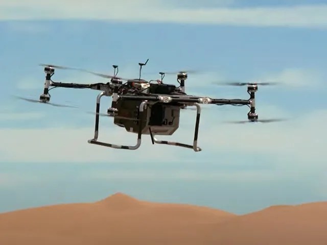 A model quadcopter is shown flying above sandy dunes with an overcast sky in the background.