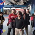 Photo of a group of 7 people smiling and interacting with a robot. The blue NASA logo is in the upper left corner.