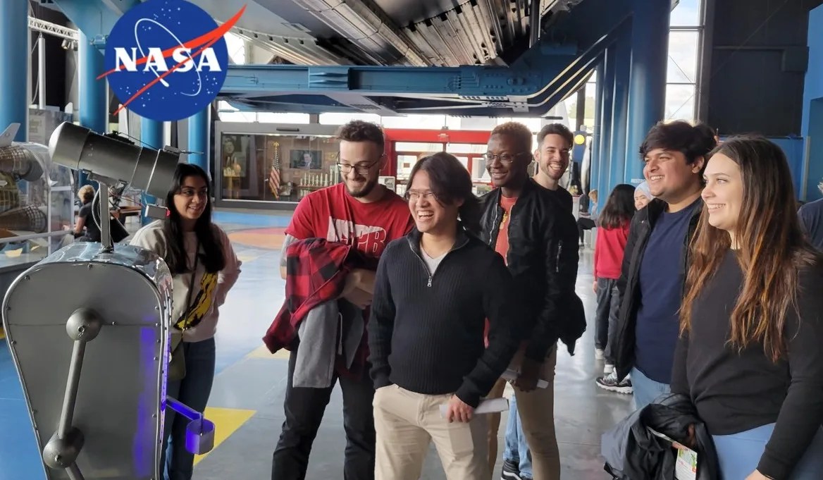 Photo of a group of 7 people smiling and interacting with a robot. The blue NASA logo is in the upper left corner.